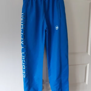 Blå träningsbyxor från Sergio Tacchini - Säljer ett par snygga blå träningsbyxor från Sergio Tacchini. De har en elastisk midja och är dekorerade med märkesnamnet längs benet. Perfekta för träning eller en avslappnad dag. Byxorna är endast använda 2 gånger och därmed i fint skick och redo för nya äventyr! XL i storlek men mer som en medium (M) i verkligheten.