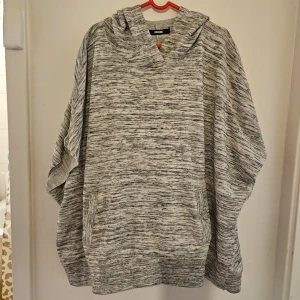 Gråmelerad poncho från Bik Bok - Säljer en gråmelerad poncho från Bik Bok med huva och fickor framtill. Perfekt för en avslappnad stil och enkel att slänga på sig när det blir kyligt. Passar bra till både jeans och leggings.