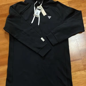 Svart hoodie från Hummel - Säljer en svart hoodie från Hummel med deras logga på bröstet. Justerbar huva med vita snören och ribbade muddar. Kan även användas som en oversized klänning för en avslappnad stil!