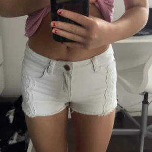 Vita shorts med spetsdetaljer - Snygga vita shorts med spetsdetaljer på sidorna. De är lågmidjade och har en knapp och dragkedja framtill. Jag säljer dom då de är små❤️