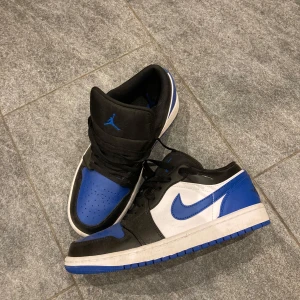 Nike Air Jordans Low 1 i svart, blå och vit - Snygga Nike Air Jordans i en cool färgkombination av svart, blå och vit. Skorna har den klassiska snörningen och den ikoniska swoosh-loggan på sidan. Perfekta för dig som vill ha en stilren och sportig look.