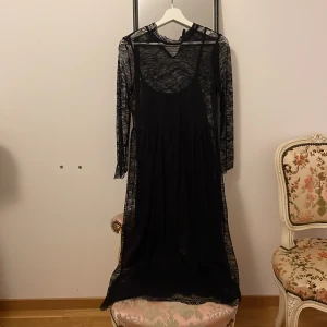 Svart spetsklänning - Elegant svart långklänning i spets med långärmad design. Klänningen har en transparent spetsöverdel och en underklänning med smala axelband. Perfekt för en stilfull kväll.