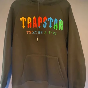 Svart hoodie från Trapstar - Snygg svart hoodie från Trapstar med färgglad text på framsidan. Den har en klassisk känguruficka och justerbar huva med dragsko. Perfekt för en avslappnad stil.