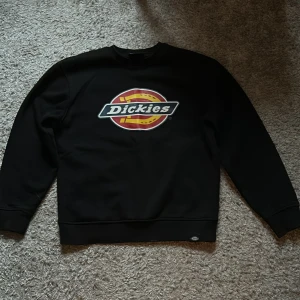 Svart sweatshirt från Dickies - Säljer en svart sweatshirt från Dickies med deras ikoniska logga i rött, gult och vitt på framsidan. Tröjan har långa ärmar och en klassisk rund halsringning. Perfekt för en avslappnad stil.