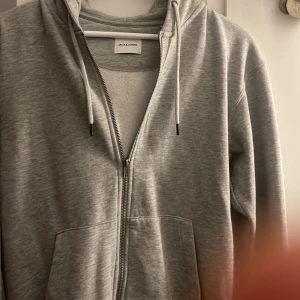 Grå hoodie från Jack & Jones - Säljer en stilren grå hoodie från Jack & Jones. Den har en dragkedja framtill och justerbar huva med snören. Perfekt för en avslappnad look.