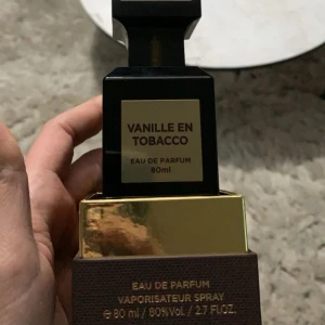 Vanille en Tobacco Eau de Parfum - En elegant flaska med Vanille en Tobacco Eau de Parfum, 80 ml. Doften kombinerar rika toner av vanilj och tobak för en sofistikerad upplevelse. Flaskan har en lyxig design med en svart och guld etikett.