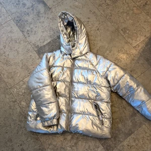 Silver pufferjacka från Stella McCartney - Snygg och glansig silverfärgad pufferjacka från Stella McCartney. Jackan har en hög krage och dragkedja framtill. Perfekt för att hålla värmen med stil under kyliga dagar.