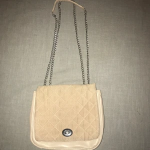 Beige quiltad axelväska med kedja - Snygg beige axelväska med quiltat mönster och silverfärgad kedja. Väskan har en praktisk stängning med ett metallspänne framtill. Perfekt för att ge en stilren touch till din outfit.