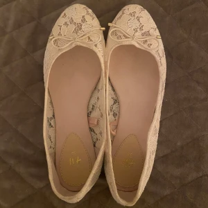 Beige/rosa spetsballerinaskor från H&M - Säljer ett par eleganta beige/rosa ballerinaskor från H&M med spetsdetaljer och en söt rosett framtill. Perfekta för en stilren look. De är helt oanvända och kommer inte till användning då dom är för små💕 