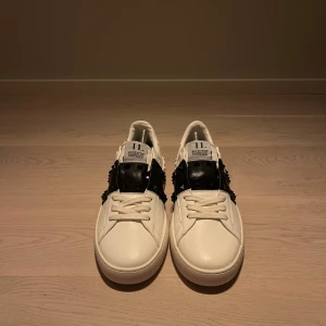 Vit och svart sneakers från Valentino - Snygga vita sneakers med svarta detaljer från Valentino. Skorna har en unik design med nitar på sidorna och en klassisk snörning. Perfekta för att ge din outfit en stilfull touch.