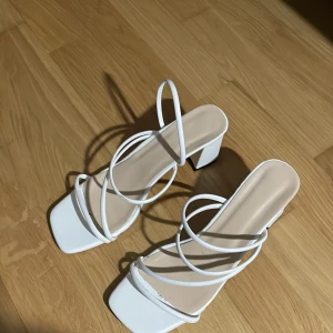 Vita sandaler med klack - Stilrena vita sandaler med klack och tunna remmar som korsar över foten. Perfekta för en elegant look med en fyrkantig tå och bekväm passform. de är andvända bara en gång på en fest men annars aldrig 