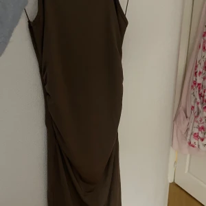 Brun klänning med knytdetalj - Elegant brun klänning med en unik knytdetalj framtill. Klänningen har en smickrande passform och är ärmlös, vilket gör den perfekt för olika tillfällen. Materialet ser ut att vara mjukt och följsamt.