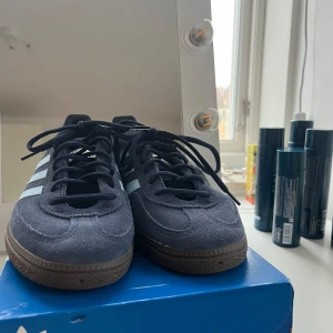 Adidas Handball Spezial sneakers - Snygga Adidas Spezial sneakers i mörkblå mocka med ljusblå detaljer och klassiska tre ränder. Skorna har en brun sula och snörning framtill. Perfekta för en stilren och sportig look.