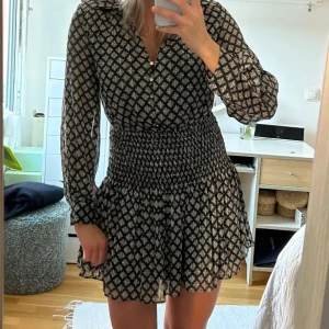 Svart zaraklänning med volang - Snygg mönstrad klänning med volangdetaljer och långa ärmar. Perfekt för både vardag och speciella tillfällen. Den har en smickrande passform och knappar framtill.