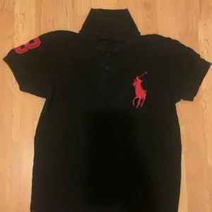 Svart Ralph louren pikétröja med röd logga - Snygg svart pikétröja från Ralph Lauren som är i fint skick, säljer den för 300kr då jag är osäker på nypriset, tveka inte att höras av vid intresse😊