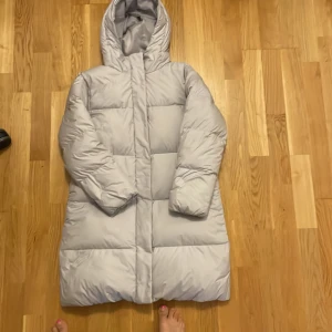 Ljusgrå dunjacka från Uniqlo - Säljer en ljusgrå dunjacka från Uniqlo med huva. Jackan är lång och har en pufferdesign som ger extra värme. Perfekt för kalla dagar.