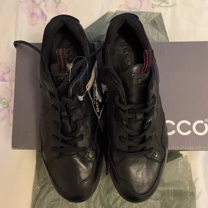 Svarta sneakers från Ecco - Snygga och praktiska svarta sneakers från Ecco med GORE-TEX teknologi för att hålla fötterna torra. Skorna har snörning och är tillverkade i skinn, vilket ger en stilren look. Perfekta för både vardag och aktivitet.