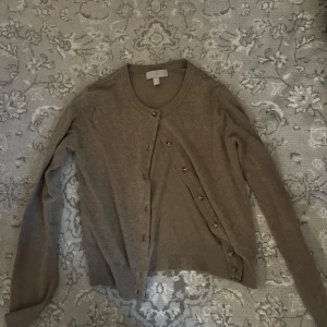 Brun kofta från H&M - Säljer en stilren brun kofta från H&M. Den har långa ärmar och knappar framtill. Perfekt för lager-på-lager under kyligare dagar.