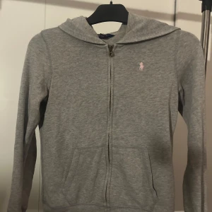 Grå hoodie från Ralph Lauren - Säljer en grå hoodie från Ralph Lauren med dragkedja och en liten rosa logga på bröstet. Perfekt för en avslappnad stil. Den har långa ärmar och en praktisk huva. Den har ett fått ett litet märke på vänstersida längst ner men inget som syns från håll.