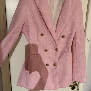 Stukturvävd figursygg blazer med gulddetaljer - Snygg rosa kavaj med dubbelknäppning och stiliga guldknappar. Perfekt för att ge en elegant touch till din outfit. Kavajen har en klassisk passform och långa ärmar. Endast använd vid fåtal tillfällen. Utmärkt skick