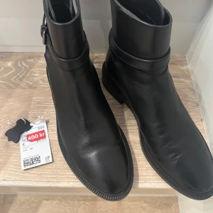 Svarta läderboots med spänne-HELT NYA - Snygga svarta boots i läder med ett stilrent spänne runt ankeln. Perfekta för både vardag och mer uppklädda tillfällen. De har en låg klack. 100% läder ytterfoder och 100% mocka inuti. Endast provade- för stora för mig. NYA! 