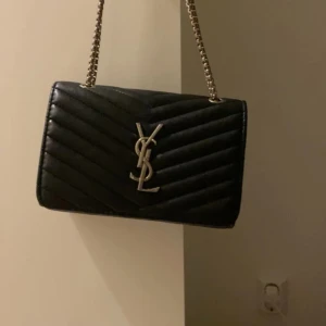 Svart quiltad axelväska från YSL - Snygg svart axelväska från YSL med quiltat mönster och kedjerem. Väskan har en elegant design med det ikoniska YSL-emblemet i silver på framsidan. Perfekt för att ge en lyxig touch till din outfit. Köpte denna väskan på plick och de finns tyvär inte bevis på att den e äkta men den ser äkta ut och materialet e bra❤️