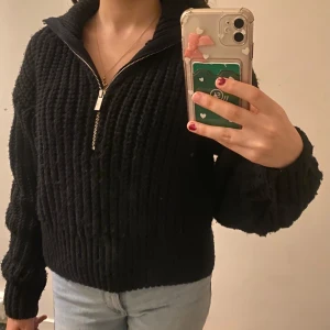 Svart stickad tröja med dragkedja - Säljer en svart stickad tröja med dragkedja och hög krage. Tröjan har långa ärmar och en ribbad struktur som ger en stilren look. Perfekt för kyligare dagar. Mina bilder!!