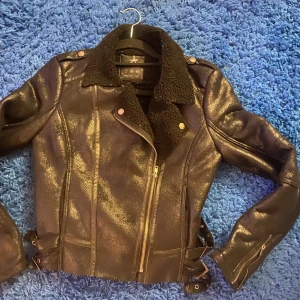 Svart skinnjacka från Atmosphere - Snygg svart skinnjacka från Atmosphere med en mjuk krage i pälsimitation. Jackan har dragkedja och spännen i midjan för en justerbar passform. Perfekt för en cool och stilren look.