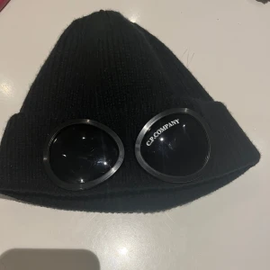 Svart mössa från C.P. Company - Snygg svart mössa från C.P. Company. Tillverkad i 100% bomull, perfekt för att hålla värmen med stil. Endast använd fåtals gånger 