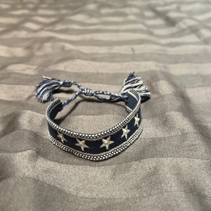 Blått armband med vita stjärnor - Snyggt blått armband med 5 stycken vita stjärnor på framsidan och flätade tofsar. Perfekt accessoar för en avslappnad stil och passar nästintill allt. Armbandet är justerbar för att passa olika storlekar. 
