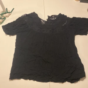 Svart blus med spetsdetaljer - Snygg svart blus med spetsdetaljer vid halsringningen. Perfekt för en stilren look. Kortärmad och i ett mjukt material. Den är inte strucken