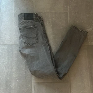 Grå jeans från Jack & Jones - Snygga grå jeans från Jack & Jones med en slim fit passform. De har en sliten look med en liten reva på ena benet. Perfekta för en avslappnad stil. Nypris 800kr ish