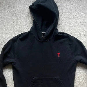 AMI Paris Hoodie Svart - Säljer en stilren svart hoodie från Ami med en röd broderad logotyp på bröstet. Tröjan har en klassisk passform med huva och dragsko. Perfekt för en avslappnad look. Storlek: M