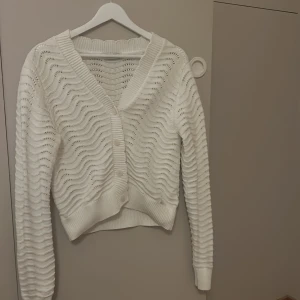 Vit stickad kofta  - Säljer en vit stickad kofta med vågigt mönster och knappar framtill. Koftan har långa ärmar och ribbade kanter, perfekt för kyliga dagar.