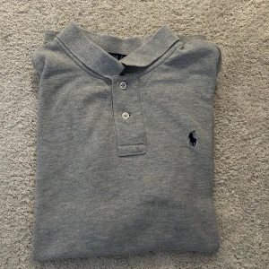 Grå pikétröja från Ralph Lauren - Säljer en klassisk grå pikétröja från Ralph Lauren med broderad logga på bröstet. Tröjan är extremt fint skick, väldigt sparsamt använt utan defekter! Storlek 14-16 alltså S. 