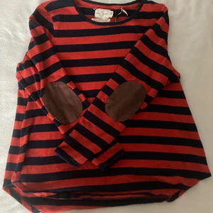 Randig tröja från Holly & Whyte - Säljer en randig tröja från Holly & Whyte by Lindex i rött och svart. Tröjan har långa ärmar och bruna armbågslappar som en snygg detalj. Perfekt för en avslappnad stil.