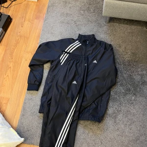 Marinblå träningsdräkt från Adidas - Säljer en marinblå träningsdräkt från Adidas med vita ränder på ärmar och byxor. Dräkten har dragkedja framtill och elastiska muddar vid ärmslut och raka neråt Perfekt för sport och fritid! Lite oversize