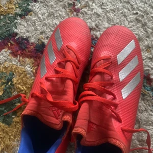 Röda fotbollsskor från Adidas - Säljer ett par röda Adidas fotbollsskor med silverfärgade detaljer och snörning. Skorna har en mönstrad yta och dobbar för bra grepp på planen. Perfekta för fotbollsträning och matcher.
