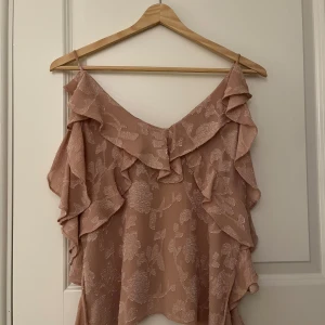 Rosa blus med volanger - Säljer en söt rosa blus med volanger. Perfekt för varma dagar och ger en romantisk touch till din outfit. Blusen har ett subtilt mönster som ger extra charm. 