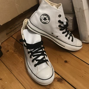 Vita Converse All Star sneakers läder  - Säljer ett par klassiska vita Converse All Star sneakers med högt skaft och svarta snören. Skorna har den ikoniska loggan på sidan och en slitstark gummisula. Perfekta för en avslappnad stil. Köpt på convers butik i Thailand för många år sedan. 