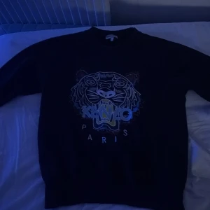 Svart tröja med tigertryck från Kenzo - Säljer en svart tröja från Kenzo med ett coolt tigertryck på framsidan. Tröjan har långa ärmar och en rund halsringning. Perfekt för en stilren och trendig look.