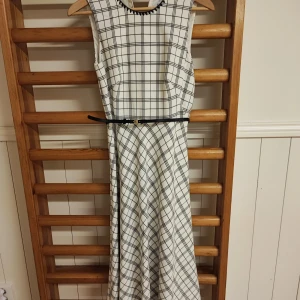 Vit och svart rutig klänning från Calvin Klein - Elegant vit och svart rutig klänning från Calvin Klein. Klänningen är ärmlös och har ett smalt bälte i midjan för en snygg silhuett. Perfekt för en stilren look.