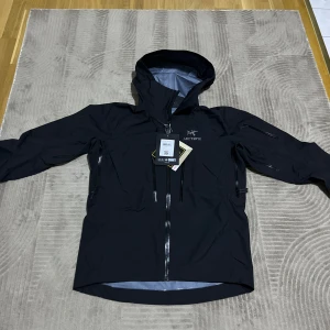 Svart skaljacka från Arc'teryx - Säljer en svart skaljacka från Arc'teryx i storlek S. Jackan är tillverkad i Gore-Tex-material som garanterar att hålla dig torr. Den har en justerbar huva och flera praktiska fickor med dragkedjor.