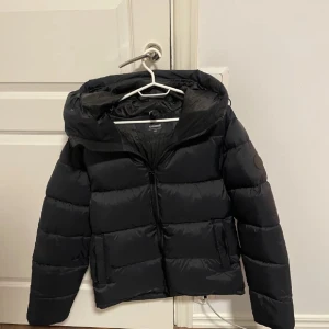 Svart pufferjacka från Everest - Säljer en svart pufferjacka från Everest med dragkedja och huva. Jackan är perfekt för kyliga dagar och har en stilren design. Den är vadderad för extra värme och komfort.