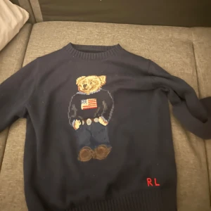 Mörkblå tröja med björnmotiv från Ralph Lauren - Säljer en mörkblå tröja från Ralph Lauren med ett broderat björnmotiv på framsidan. Tröjan har rund hals och långa ärmar. Perfekt för en avslappnad stil med en touch av lekfullhet.
