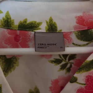 Blommig klänning från Vero Moda - Säljer en söt blommig byxdress från Vero Moda i storlek XS. Byxdressen har ett vackert blommönster i rosa och grönt på en vit bakgrund. Den har korta ärmar och en omlott-design som ger en fin passform. Perfekt för sommarens alla tillfällen! 🌸