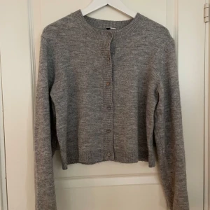 Grå stickad kofta - Säljer en mysig grå stickad kofta med knappar framtill. Perfekt för kyligare dagar och enkel att matcha med olika outfits. Koftan har långa ärmar och en klassisk rund halsringning.