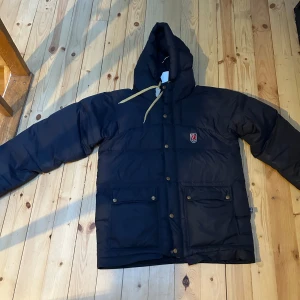 Mörkblå dunjacka från Fjällräven - Säljer en mörkblå dunjacka från Fjällräven med huva och knäppning framtill. Jackan har två stora fickor med knappar och en liten logotyp på bröstet. Perfekt för kyliga dagar!