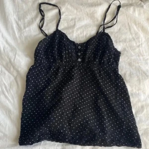Svart prickig linnetopp - Säljer en söt svart linnetopp med vita prickar och smala axelband. Toppen har knappar framtill och en lätt rynkad detalj under bysten för en smickrande passform. Perfekt för varma dagar! finns ingen storlek men uppskappae XS