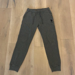 Grå mjukisbyxor från USPA Sport - Snygga grå mjukisbyxor från USPA Sport med elastisk midja och snörning. Byxorna har en broderad logga på sidan och muddar vid bensluten för en bekväm passform. Snygga och sköna mjukisbyxor, nypris 450kr. Bra pris!